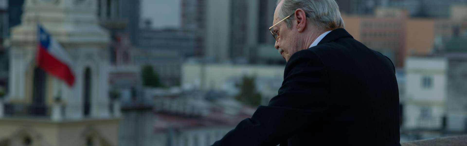Still from Wild Horse Nine: an older man in a dark suit leans on a balcony wall overlooking a city, with a red, white and blue flag on a rooftop.