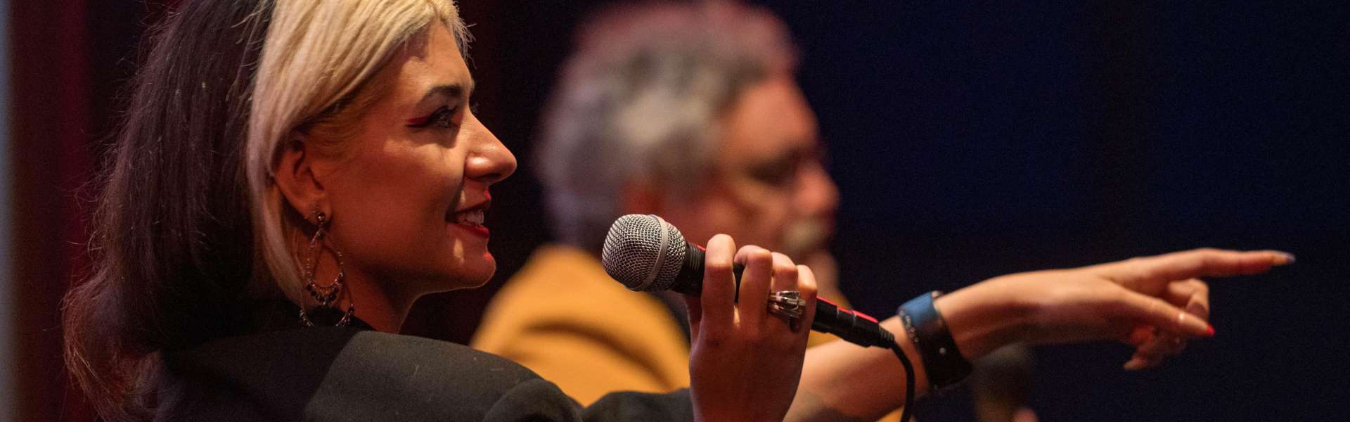 A woman with silver-blonde hair holds a microphone and points to someone off-camera during a talk, with a man seated behind her.