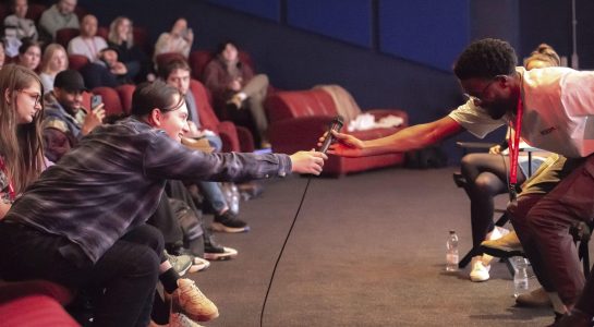 A cinema audience looks on as a man in the aisle reaches out to a person in the front row.