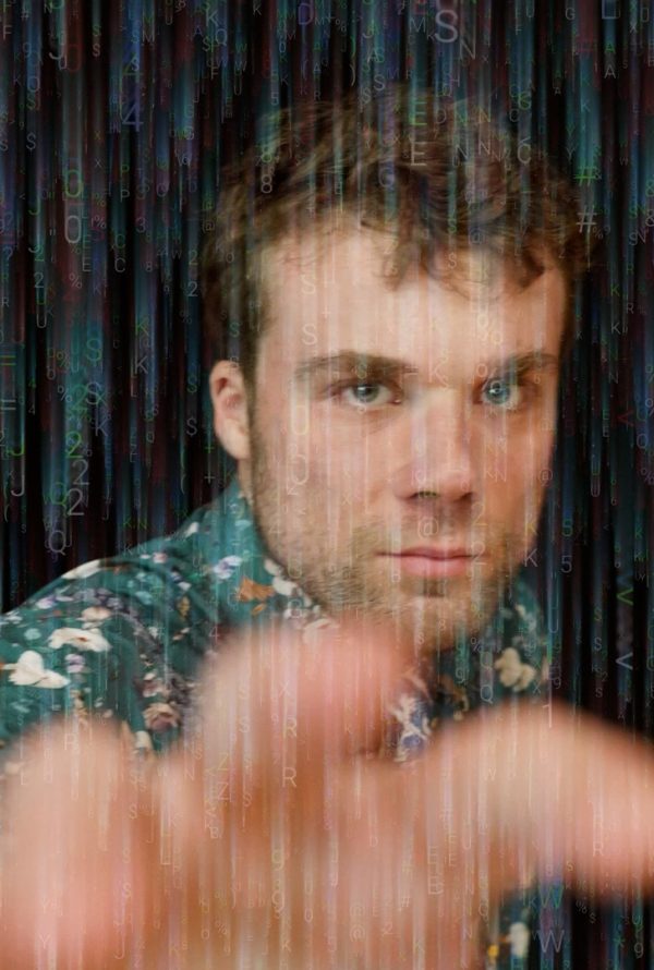 Still from Cambridge Connections: a man in a patterned shirt reaches a hand towards the camera against a dark background streaked with blue and green light.