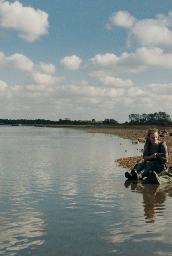 Still from Green Grass: two people sit on the shore of a calm estuary under a blue sky with clouds.