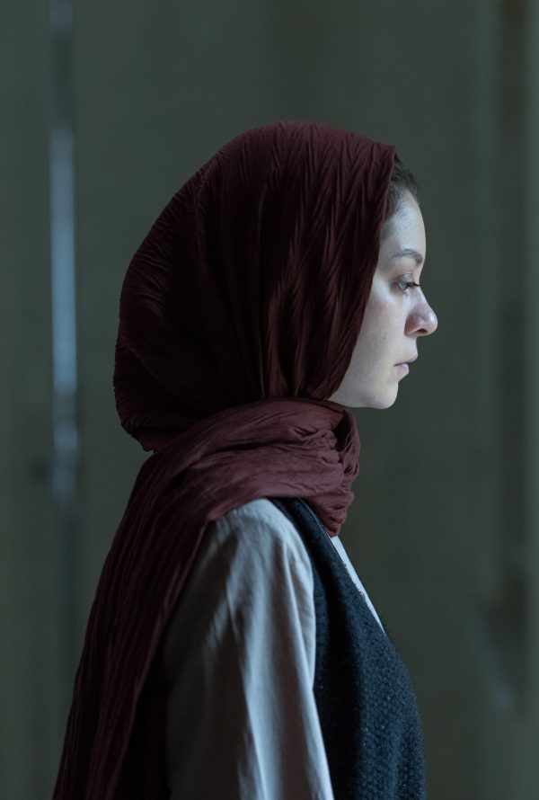 Still from It Means Hope: a woman in a red headscarf seen in profile in a dim interior.