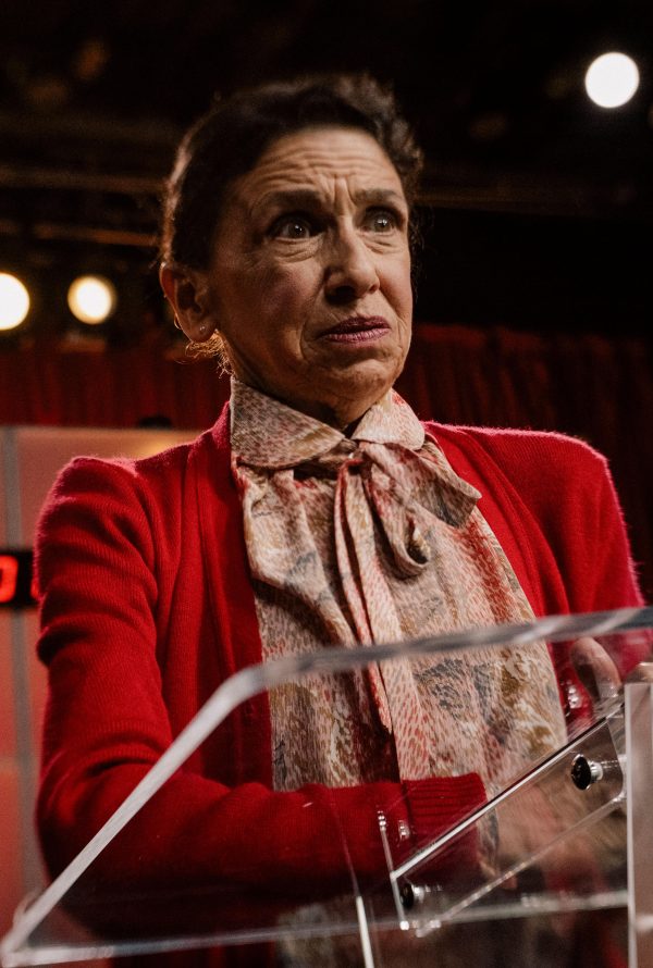 Still from Jasmine. Home. Mother.: a woman in a red jacket and ruffled cream blouse stands at a clear podium under bright stage lights, looking to one side with a tense expression.