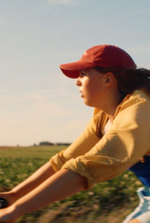 Still from The Harvest: a girl in a red cap and yellow top rides a bicycle fast across a field, her hair blowing back.